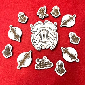 Puede incluir: Un conjunto de piezas de juego de madera cortadas con láser sobre una superficie roja. Las piezas incluyen un escudo central con las letras "RDP" y el número "10", junto con otras formas como espadas, calaveras y un águila.
