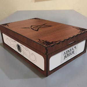 Puede incluir: Una caja de almacenamiento rectangular de madera con un acabado marrón oscuro. La tapa presenta un símbolo de estrella grabado con láser y diseños en las esquinas. Los paneles laterales son blancos con ilustraciones en tinta negra de tentáculos y el texto Arkham Horror.