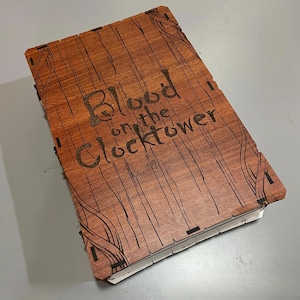 Puede incluir: Una caja de madera con el texto "Blood on the Clocktower" grabado en la tapa. La caja tiene un patrón de vetas de madera y diseños decorativos en las esquinas. La caja está cerrada y parece estar sobre una superficie gris.