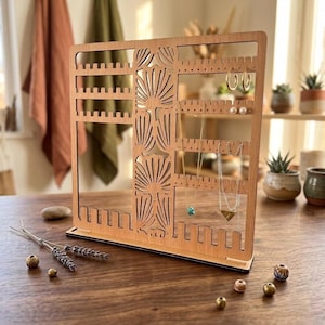 Könnte beinhalten: Ein Holz-Schmuck-Organizer mit einem lasergeschnittenen Blumendesign. Der Organizer hat mehrere Schlitze und Löcher für Ohrringe, Halsketten und anderen Schmuck. Die natürliche Holzfarbe ergänzt den ausgestellten Schmuck.