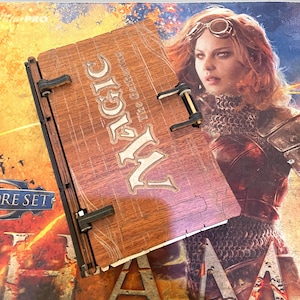 May include: Wooden Magic: The Gathering card holder with the word "MAGIC" in large, stylized letters. The holder has a distressed wood grain appearance and black metal hinges. The background features a woman with goggles and a Core Set box.
