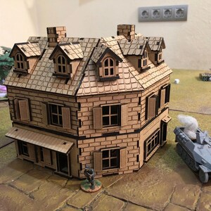 May include: A detailed, two-story model building with a brick facade, dormer windows, and a tiled roof. The building has shutters on the windows and a small awning. A miniature military vehicle is parked nearby, suggesting a war game setting.