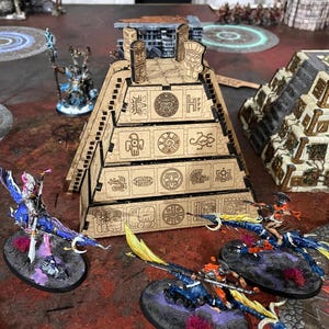 Puede incluir: Una escena de juego de mesa con una pirámide de madera cortada con láser y figuras en miniatura. La pirámide es beige con símbolos grabados. Varias figuras de fantasía con alas azules y moradas y jinetes están posicionadas alrededor.