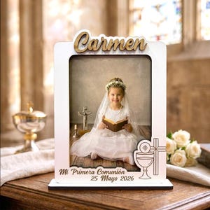 Puede incluir: Un marco de fotos de madera blanca con el nombre "Carmen" en fuente de madera en la parte superior. El marco contiene una foto de una niña con un vestido blanco. El texto "Mi Primera Comunión 25 Mayo 2026" está en la parte inferior.