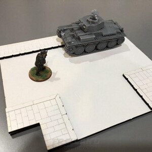 Może przedstawiać: Szary miniaturowy czołg ze szczegółowymi gąsienicami i wieżą stoi na białej powierzchni. Miniaturowy żołnierz stoi w pobliżu na okrągłej podstawie z zielonymi detalami. Scena rozgrywa się na białej podstawie z wzorem cegieł.