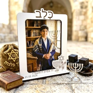 May include: A white wooden picture frame with Hebrew text at the top and bottom, holding a photo of a young person in a blue suit and prayer shawl. A menorah and other religious items are displayed.