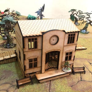May include: A miniature, two-story building with a clock on the facade, designed for tabletop gaming. The building is made of light brown wood with a tiled roof and small benches. A miniature figure stands on the porch.