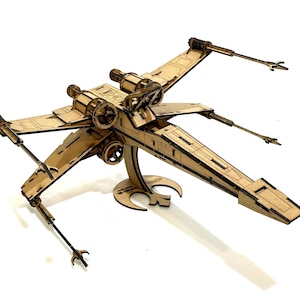 Peut inclure: Maquette en bois d'un X-wing, un vaisseau spatial de la franchise Star Wars. Le modèle est en bois brun clair avec des détails complexes, notamment des ailes, des moteurs et un support. Le modèle mesure environ 30 cm de long.
