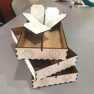 Puede incluir: Una pila de cajas de almacenamiento de madera cortadas con láser con símbolos grabados. Las cajas son de color beige con bordes y divisores de color marrón oscuro. Los símbolos incluyen una llama, una gota de agua, un sol, un árbol, una calavera y un diamante.