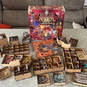 Puede incluir: Un juego de mesa con una caja grande con ilustraciones de fantasía coloridas, rodeado de contenedores de almacenamiento de madera llenos de piezas de juego, cartas y fichas. Los componentes del juego están dispuestos sobre una mesa.