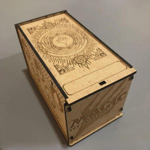 Puede incluir: Una caja de madera de color marrón claro con diseños grabados con láser y el texto "MAGIC THE GATHERING". La caja es rectangular con una tapa con bisagras. La parte superior de la caja presenta un diseño circular.