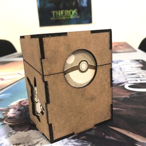 Puede incluir: Una caja de madera marrón con un diseño circular que se asemeja a una Poké Ball en la parte delantera. Un recorte de un personaje de dibujos animados es visible en el lateral. La caja está diseñada para guardar cartas coleccionables o pequeños objetos.