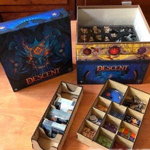 May include: A board game box set with the word "DESCENT" in orange lettering. The box is dark blue with a detailed illustration. Several wooden storage trays are visible, filled with game pieces and cards.