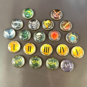 Op de afbeelding: Een verzameling van eenentwintig ronde speltoken met verschillende ontwerpen. Sommige tokens tonen afbeeldingen van een stier, een demon, een schedel en een vlam. Andere tonen Romeinse cijfers I tot en met V. Verschillende tokens hebben de tekst "+1/+1" en "-1/-1".