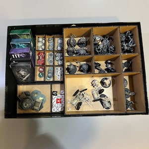 Pode incluir: Uma caixa de jogo preta com vários compartimentos cheios de peças de jogo, incluindo figuras em miniatura, dados e fichas de cartão. A caixa é organizada com divisórias de madeira.
