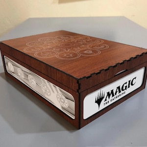 Puede incluir: Una caja de madera marrón oscuro con un diseño grabado en la tapa. La caja presenta un panel blanco con el logotipo "MAGIC: THE GATHERING". El panel lateral tiene un diseño plateado detallado.