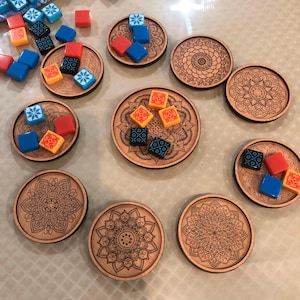 Puede incluir: Posavasos de madera con intrincados diseños de mandala. Cada posavasos contiene piezas de juego de colores: rojo, azul, naranja y negro. Los posavasos están dispuestos sobre una superficie de color claro, creando una disposición visualmente atractiva.