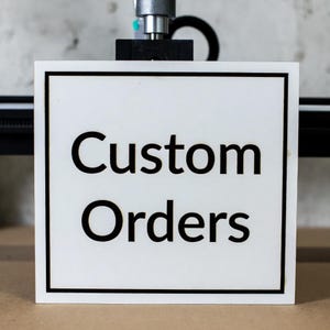 Puede incluir: Un letrero cuadrado blanco con un borde negro muestra las palabras "Pedidos personalizados" en negrita, en negro. El letrero está montado en una máquina, lo que sugiere un servicio de fabricación o personalización.
