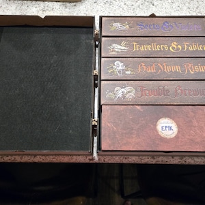 Peut inclure: Boîte en bois ouverte avec un intérieur vert foncé et une série de boîtes marron. Les boîtes sont étiquetées avec du texte, notamment "Sects & Violets", "Travellers & Fabled", "Bad Moon Rising" et "Trouble Brewing". La boîte inférieure porte un autocollant circulaire avec le texte "EPIK".