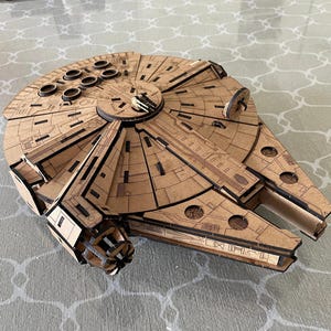 May include: A detailed, laser-cut wooden model of the Millennium Falcon starship. The model features intricate paneling and circular details, with a light brown color and dark brown accents. The iconic spaceship is a popular item for Star Wars fans.