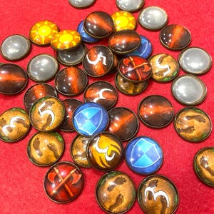 Puede incluir: Una colección de fichas de juego redondas con varios diseños. Algunas fichas presentan un sol, el número 5, huellas, hachas cruzadas y un escudo. Las fichas tienen un borde metálico y están sobre un fondo rojo.