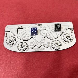 Könnte beinhalten: Ein silberfarbenes Spielbrett mit dem Wort "TURN" und dem Text "C.P." aufgedruckt. Zwei Würfel, einer blau und einer schwarz, liegen auf dem Brett. Das Brett hat Zifferblätter mit der Aufschrift "PRIMARY" und "SECONDARY".