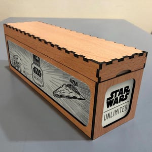 Può includere: Scatola rettangolare in legno con dettagli tagliati al laser e la scritta "STAR WARS UNLIMITED". La scatola presenta grafiche di Star Wars, tra cui un Star Destroyer. Realizzata in legno chiaro con dettagli neri.
