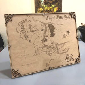 Puede incluir: Un mapa de madera de la Tierra Media de El Señor de los Anillos, con intrincados detalles cortados con láser. El mapa es de color marrón claro, con detalles decorativos en las esquinas. El texto "Map of Middle Earth" y "The Lord of the Rings" son visibles.