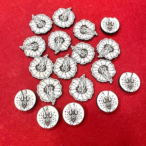 May include: A collection of wooden game pieces on a red surface. The pieces include circular tokens with numbers and a horned figure, and floral-shaped tokens with flame designs. The tokens are black and white.
