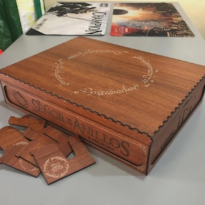 May include: A wooden box with laser-etched designs and the text "SEÑOR DE LOS ANILLOS" in a decorative font. The box is brown and has a rectangular shape. Several wooden game pieces are next to the box.