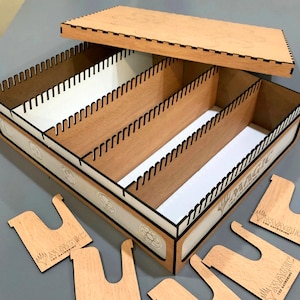 May include: A wooden card storage box with a lid, featuring multiple compartments. The box has a light brown finish with black trim and white interior dividers. The words "MAGIC THE GATHERING" are visible on the side and on the included dividers.