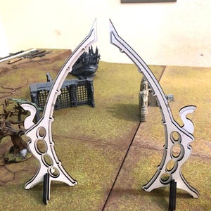 Könnte beinhalten: Zwei weiß-schwarze dekorative Torbögen mit geschwungenem Design und ovalen Aussparungen. Die Torbögen stehen auf einer grün-braunen Oberfläche, mit Miniaturgebäuden im Hintergrund. Die Torbögen sind wahrscheinlich für ein Tabletop-Spiel.