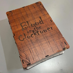 May include: A wooden box with the text "Blood on the Clocktower" engraved on the lid. The box has a wood grain pattern and decorative corner designs. The box is rectangular and appears to be a game box.