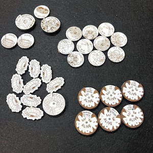 May include: A collection of circular and irregular game tokens featuring engraved illustrations and text. The tokens include designs of creatures, bones, and text such as WARD, EAT 'EM ALIVE, and various game mechanics. The tokens are white with brown details.