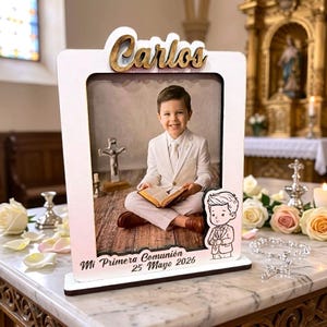 May include: A white and wood-toned photo frame with the name "Carlos" in gold script. The frame holds a photo of a smiling boy in a white suit, holding a book. The text "Mi Primera Comunión 25 Mayo 2026" is at the bottom.