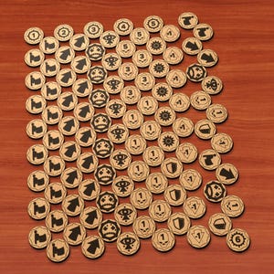 Puede incluir: Una colección de fichas de juego circulares de madera con varios iconos negros grabados, incluyendo los números del uno al seis, flechas, calaveras, ojos, escudos y figuras sosteniendo banderas, dispuestos en un patrón de cuadrícula denso.