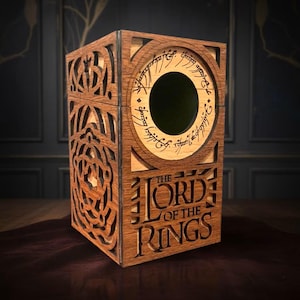 Puede incluir: Caja de madera con intrincados diseños calados y las palabras "THE LORD OF THE RINGS" talladas en la parte delantera. La caja presenta un diseño circular con escritura alrededor de un agujero central. La madera tiene un tono cálido y natural.