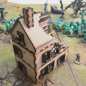 May include: A miniature, two-story, laser-cut wooden house with a distressed appearance. The roof and walls are partially damaged, with visible window frames and a chimney. The house is set on a terrain board with miniature figures, suggesting a tabletop game setting.