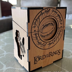 May include: A wooden box with laser-cut designs from "The Lord of the Rings." The box features a circular design with the inscription and a hand-shaped cutout. The words "THE LORD OF THE RINGS" are displayed on the front.
