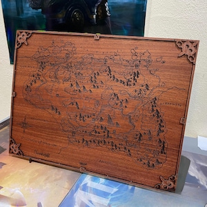 Puede incluir: Un mapa de madera detallado de un mundo de fantasía, con montañas, ríos y ciudades. El mapa está enmarcado con esquinas decorativas e indicadores direccionales. La madera es de color marrón rojizo.