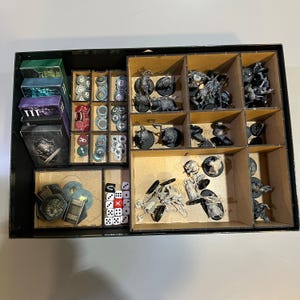 Pode incluir: Uma caixa de jogo preta com vários compartimentos cheios de peças de jogo, incluindo figuras em miniatura, dados e fichas de cartão. A caixa é organizada com divisórias de madeira, e as peças do jogo são de várias cores.