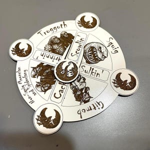 Puede incluir: Una pieza de juego circular de color crema con un puntero giratorio. La pieza está dividida en secciones con ilustraciones y texto, incluyendo "Troggoth", "Squig" y "Cack". Formas decorativas de media luna se colocan alrededor del perímetro.