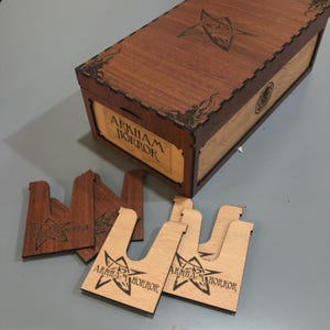 a wooden box with the label "AARKHAM HORROR" and several wooden pieces with the same label, likely cutouts or cards, placed on a gray surface.