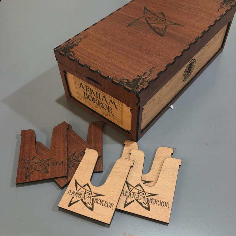 a wooden box with the label "AARKHAM HORROR" and several wooden pieces with the same label, likely cutouts or cards, placed on a gray surface.