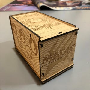 Puede incluir: Caja de madera clara con diseños grabados y la inscripción "MAGIC THE GATHERING". La caja rectangular tiene una tapa. El lateral de la caja tiene la palabra "COMMANDER" y otros símbolos grabados.
