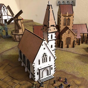Op de afbeelding: Een miniatuurmodel van een witte kerk met een bruin dak en een hoge toren. Andere gebouwen, waaronder een windmolen, staan op de achtergrond. De scène speelt zich af op een groen en bruin oppervlak, met kleine beeldjes op de voorgrond.