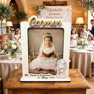 Puede incluir: Un marco con una foto de una niña con un vestido blanco, velo y corona de flores, sosteniendo un libro. El marco tiene el nombre "Carmen" en letras doradas y el texto "Mi Primera Comunión 25 Mayo 2026". Una ilustración de dibujos animados de una niña está en el marco.