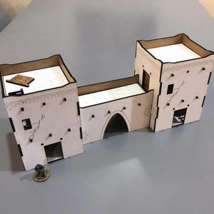 May include: A laser-cut, unpainted wooden model of a fortress or town, featuring two towers connected by a wall with an arched gateway. The model has detailed cutouts for windows and doors, and decorative patterns along the edges.