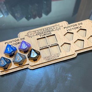 May include: Wooden Warhammer Age of Sigmar game board with dice. The board has slots for dice and text in Spanish. Six multi-sided dice are in the image, with numbers 1, 2, 3, 5, 7, and 9 visible.