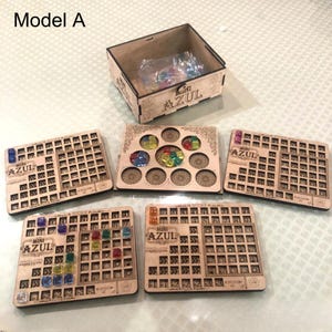 Puede incluir: Un juego de mesa Mini Azul de madera, que incluye una caja y varias tableros con patrones grabados y el logotipo "Mini Azul". Los tableros tienen espacios para piezas de juego coloridas. El juego se muestra sobre una superficie clara.